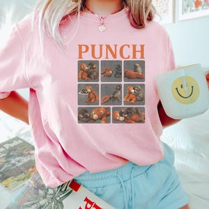 May include: Pink sweatshirt with the word "PUNCH" in orange block letters above a grid of teddy bear illustrations. The sweatshirt is paired with light blue shorts. A white mug with a yellow smiley face is held in the hand.