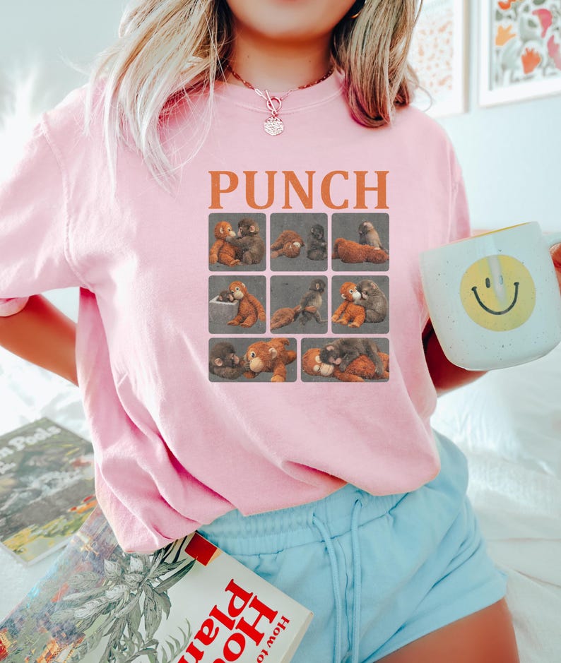 May include: Pink sweatshirt with the word "PUNCH" in orange block letters above a grid of teddy bear illustrations. The sweatshirt is paired with light blue shorts. A white mug with a yellow smiley face is held in the hand.
