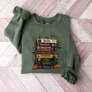 May include: An olive green crewneck sweatshirt featuring a graphic print of a stack of VHS tapes titled Addams Family, Hocus Pocus, Beetlejuice, Halloweentown, Halloweentown II, The Nightmare Before Christmas, Goosebumps, and The Witches with pumpkins.