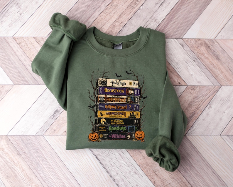 May include: An olive green crewneck sweatshirt featuring a graphic print of a stack of VHS tapes titled Addams Family, Hocus Pocus, Beetlejuice, Halloweentown, Halloweentown II, The Nightmare Before Christmas, Goosebumps, and The Witches with pumpkins.