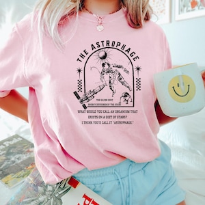May include: Pink t-shirt with a black graphic of an astronaut, sun, and text that reads "THE ASTROPHAGE" and "ENERGY DEVOURER OF THE STARS." The shirt also has the text "WHAT WOULD YOU CALL AN ORGANISM THAT EXISTS ON A DIET OF STARS? I THINK YOU'D CALL IT 'ASTROPHAGE.'"