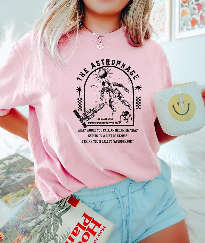 May include: Pink t-shirt with a black graphic of an astronaut, sun, and text that reads "THE ASTROPHAGE" and "ENERGY DEVOURER OF THE STARS." The shirt also has the text "WHAT WOULD YOU CALL AN ORGANISM THAT EXISTS ON A DIET OF STARS? I THINK YOU'D CALL IT 'ASTROPHAGE.'"