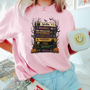 May include: A light pink t-shirt featuring a graphic print of a stack of VHS tapes titled The Addams Family, Hocus Pocus, Beetlejuice, Halloweentown, Halloweentown II, The Nightmare Before Christmas, Goosebumps, and The Witches, framed by bare tree branches.