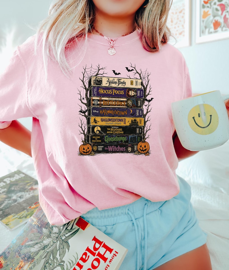May include: A light pink t-shirt featuring a graphic print of a stack of VHS tapes titled The Addams Family, Hocus Pocus, Beetlejuice, Halloweentown, Halloweentown II, The Nightmare Before Christmas, Goosebumps, and The Witches, framed by bare tree branches.
