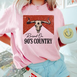 May include: A light pink sweatshirt with the text "Raised On 90's Country" and a graphic of a bull skull. The graphic is in a rectangular shape with a red background. The sweatshirt is paired with light blue shorts.
