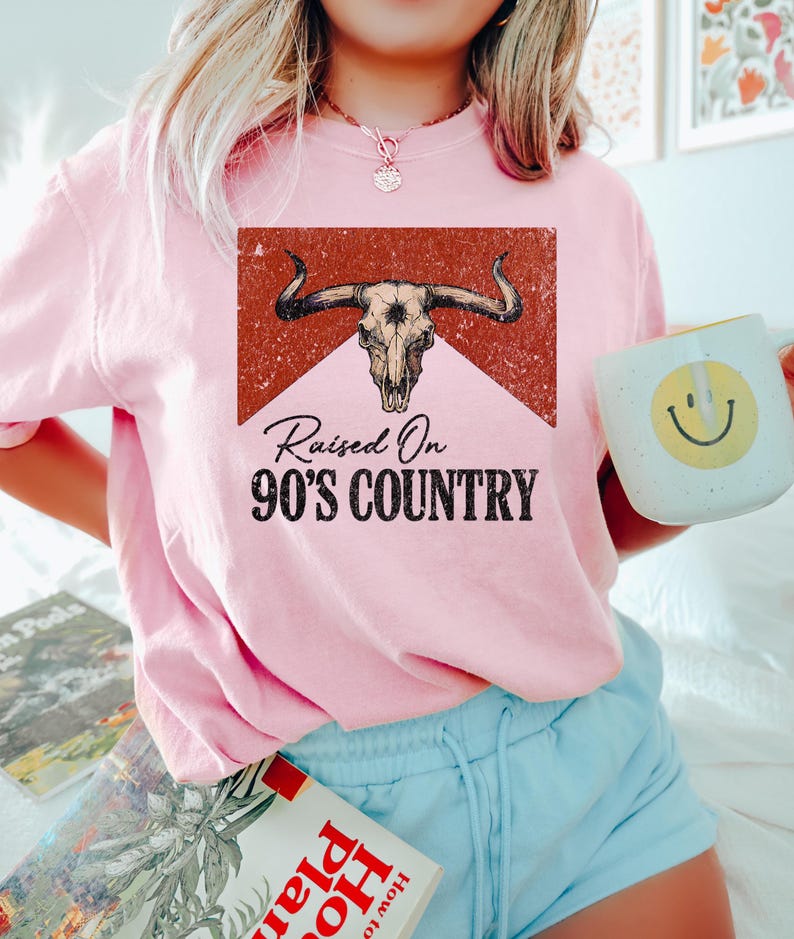 May include: A light pink sweatshirt with the text "Raised On 90's Country" and a graphic of a bull skull. The graphic is in a rectangular shape with a red background. The sweatshirt is paired with light blue shorts.