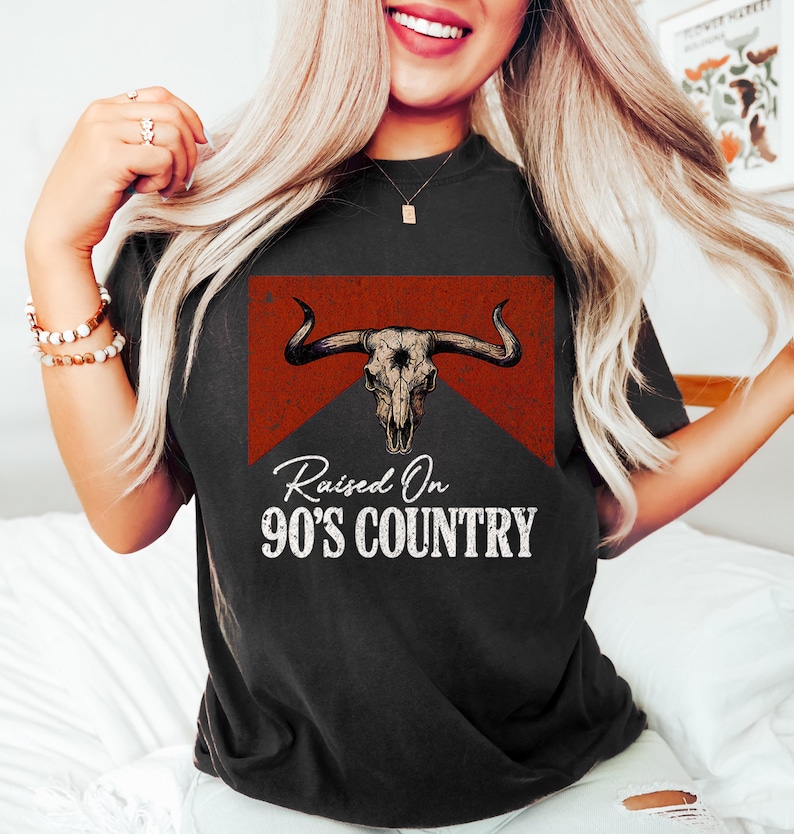 May include: Black t-shirt featuring a steer skull graphic with the text "Raised On 90's Country" in white. The graphic is set against a red and black background. The shirt is a casual wear item.