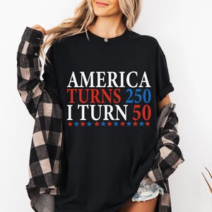 May include: Black t-shirt with the text "AMERICA TURNS 250 I TURN 50" in white, red, and blue. The design includes red and blue stars. The person is wearing a black and brown plaid shirt over the t-shirt.