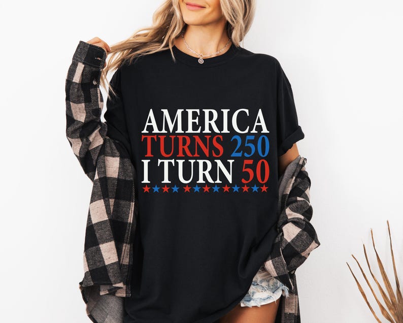 May include: Black t-shirt with the text "AMERICA TURNS 250 I TURN 50" in white, red, and blue. The design includes red and blue stars. The person is wearing a black and brown plaid shirt over the t-shirt.