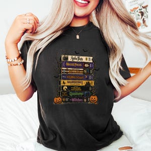May include: A black t-shirt featuring a graphic print of a stack of vintage VHS tapes. The titles include The Addams Family, Hocus Pocus, Beetlejuice, Halloweentown, Halloweentown II, The Nightmare Before Christmas, Goosebumps, and The Witches.