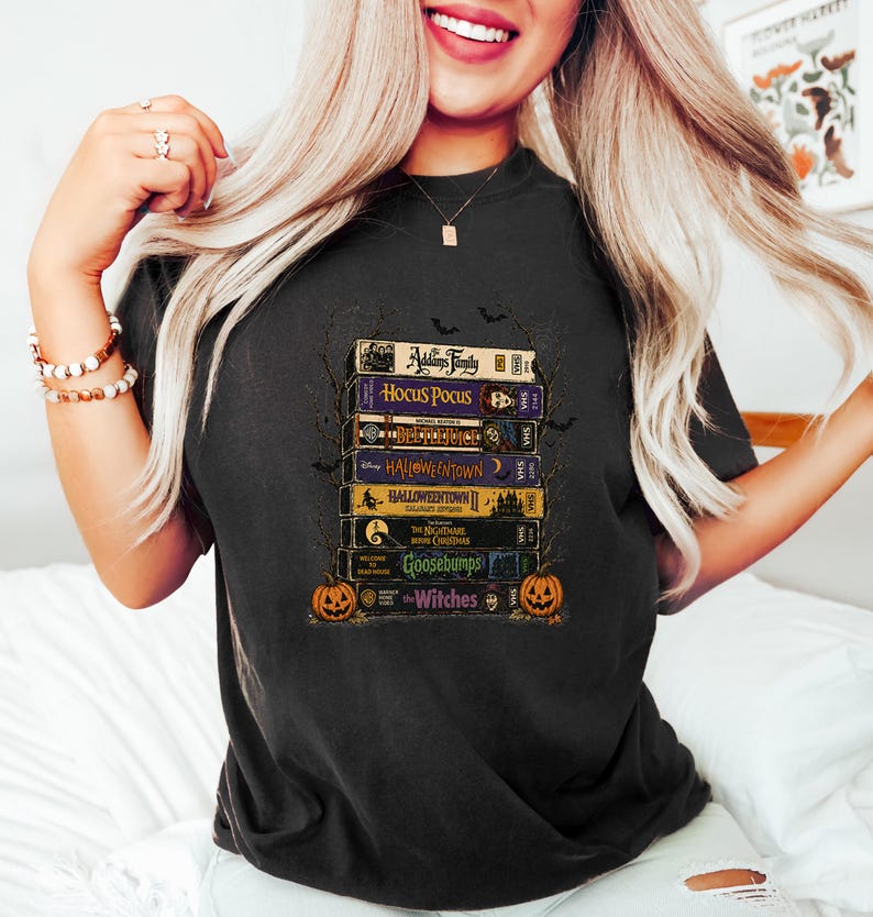 May include: A black t-shirt featuring a graphic print of a stack of vintage VHS tapes. The titles include The Addams Family, Hocus Pocus, Beetlejuice, Halloweentown, Halloweentown II, The Nightmare Before Christmas, Goosebumps, and The Witches.