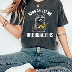May include: A charcoal gray short-sleeve t-shirt featuring a graphic of a raccoon holding a yellow measuring tape. The text above the raccoon reads HANG ON, LET ME and the text below reads OVER-ENGINEER THIS in white capital letters.