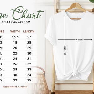 May include: A white Bella Canvas 3001 t-shirt displayed on a wooden hanger, with a size chart to the left. The chart shows measurements in inches for sizes XS to 5XL. The shirt's width and length are indicated with arrows.
