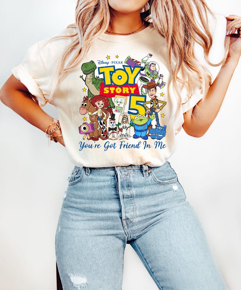 May include: A cream-colored short-sleeve t-shirt featuring a colorful graphic print of Disney Pixar Toy Story characters including Woody, Buzz Lightyear, Jessie, and Rex. The text reads Toy Story 5 and You've Got Friend In Me with yellow stars.