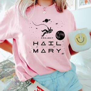 May include: Pale pink sweatshirt featuring a black space-themed graphic with an astronaut, planets, and stars. The text "PROJECT HAIL MARY" is printed below. A white mug with a yellow smiley face is held.