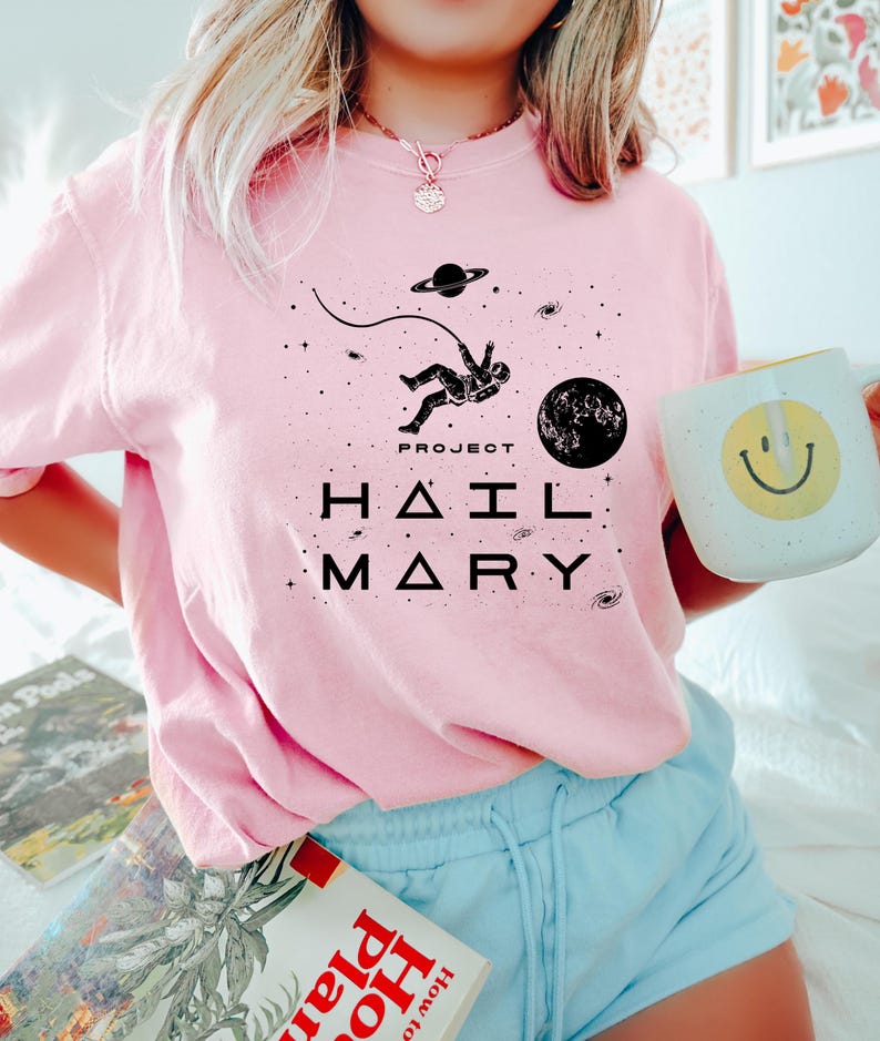May include: Pale pink sweatshirt featuring a black space-themed graphic with an astronaut, planets, and stars. The text "PROJECT HAIL MARY" is printed below. A white mug with a yellow smiley face is held.