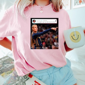 May include: A light pink t-shirt featuring a graphic print of a social media post. The graphic shows a person in a dark blue basketball jersey pointing, with the text "SOPHIE Following" and "I'm so glad I got my nails done for this" above the image.