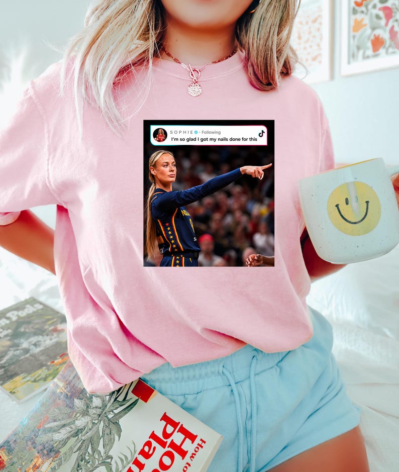 May include: A light pink t-shirt featuring a graphic print of a social media post. The graphic shows a person in a dark blue basketball jersey pointing, with the text "SOPHIE Following" and "I'm so glad I got my nails done for this" above the image.
