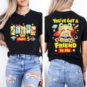 May include: A black short-sleeve t-shirt featuring Toy Story 5 graphics. The front displays characters in rectangular frames with the text Toy Story 5. The back shows characters watching a screen with the text You've Got A Friend In Me.