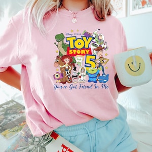 May include: A light pink t-shirt featuring a colorful graphic of Disney Pixar Toy Story characters including Woody, Buzz Lightyear, Jessie, Rex, and others, with the text Toy Story 5 and You've Got Friend In Me printed in blue and yellow.