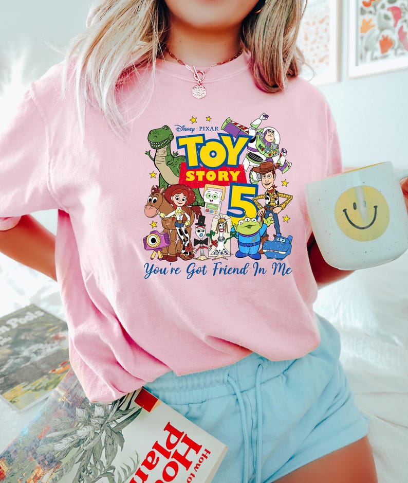 May include: A light pink t-shirt featuring a colorful graphic of Disney Pixar Toy Story characters including Woody, Buzz Lightyear, Jessie, Rex, and others, with the text Toy Story 5 and You've Got Friend In Me printed in blue and yellow.