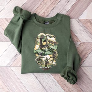 Puede incluir: Sudadera verde de cuello redondo con un gr&aacute;fico de la serie Star Wars, "The Mandalorian and Grogu". El dise&ntilde;o incluye im&aacute;genes del casco del Mandaloriano, Grogu y otros personajes. La frase "This is the Way" est&aacute; impresa debajo.