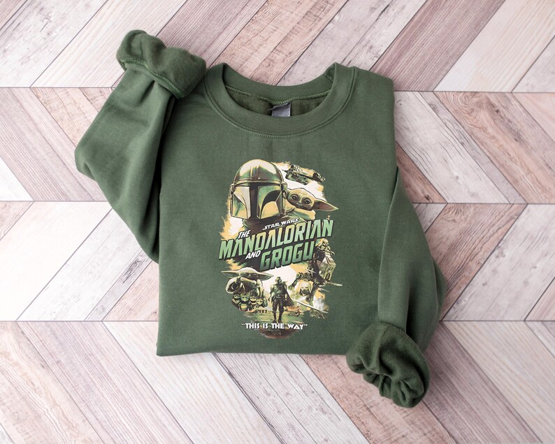 Puede incluir: Sudadera verde de cuello redondo con un gr&aacute;fico de la serie Star Wars, "The Mandalorian and Grogu". El dise&ntilde;o incluye im&aacute;genes del casco del Mandaloriano, Grogu y otros personajes. La frase "This is the Way" est&aacute; impresa debajo.