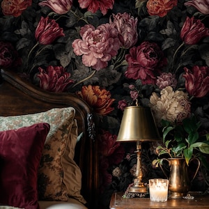 May include: A bedroom scene featuring a dark floral wallpaper with large blooms in shades of red, pink, and cream. A wooden bed frame, pillows, a bedside table with a lamp, candle, and a brass pitcher with greenery complete the cozy setting.