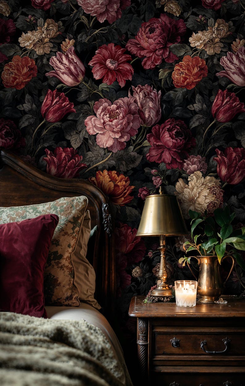 May include: A bedroom scene featuring a dark floral wallpaper with large blooms in shades of red, pink, and cream. A wooden bed frame, pillows, a bedside table with a lamp, candle, and a brass pitcher with greenery complete the cozy setting.