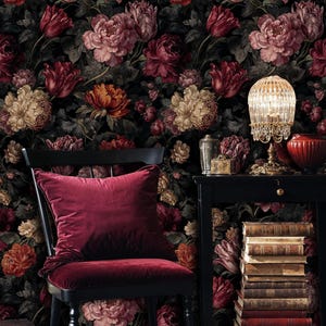 May include: A dark, floral wallpaper backdrop featuring a variety of flowers in shades of red, pink, and cream. A black chair with a burgundy velvet cushion and seat is in the foreground. A small table with a lamp and books is to the right.