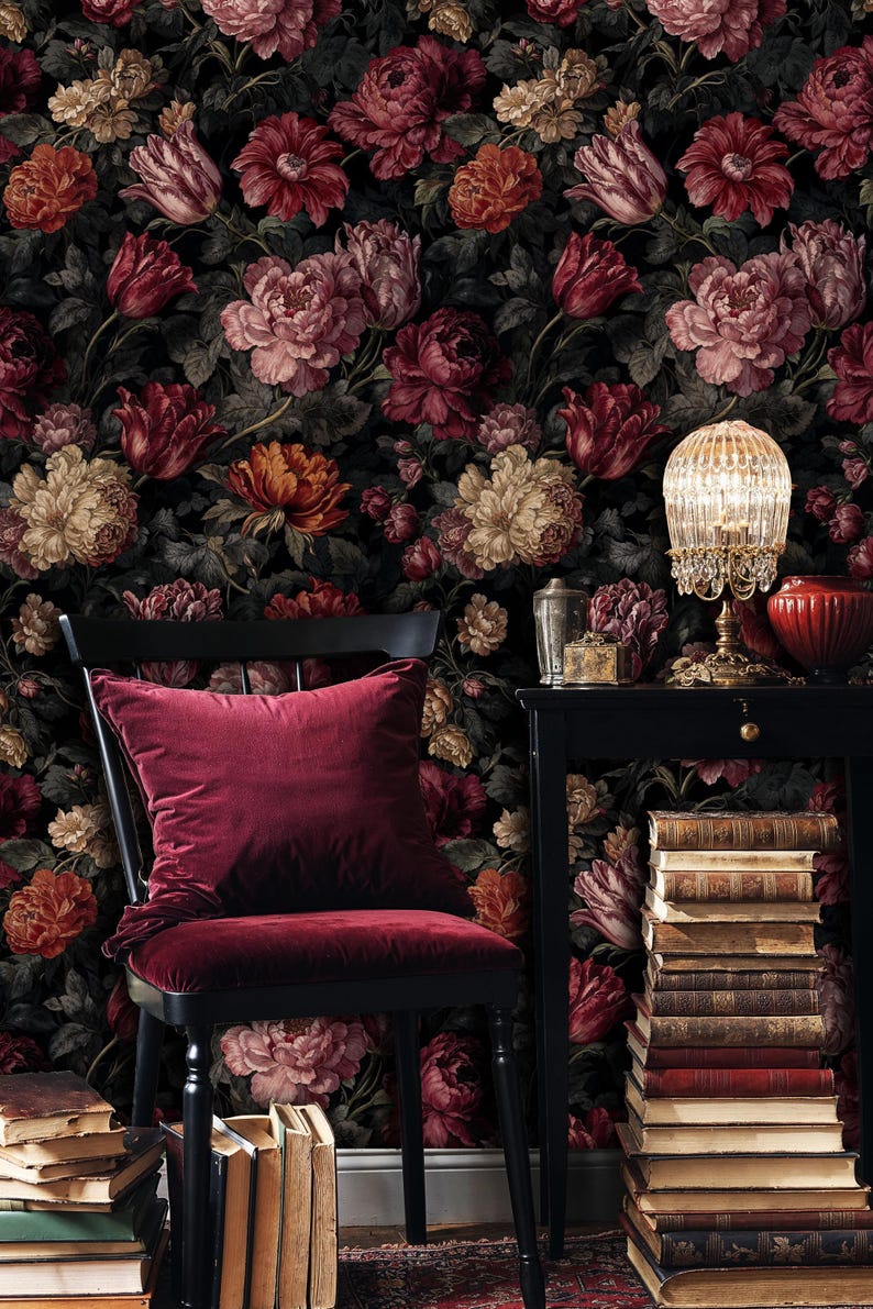 May include: A dark, floral wallpaper backdrop featuring a variety of flowers in shades of red, pink, and cream. A black chair with a burgundy velvet cushion and seat is in the foreground. A small table with a lamp and books is to the right.