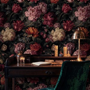 May include: A dark wooden desk with a brass lamp, books, and candles sits in front of a floral wallpaper. A green velvet chair is in the foreground. The wallpaper features large, colorful flowers in shades of red, pink, and orange.