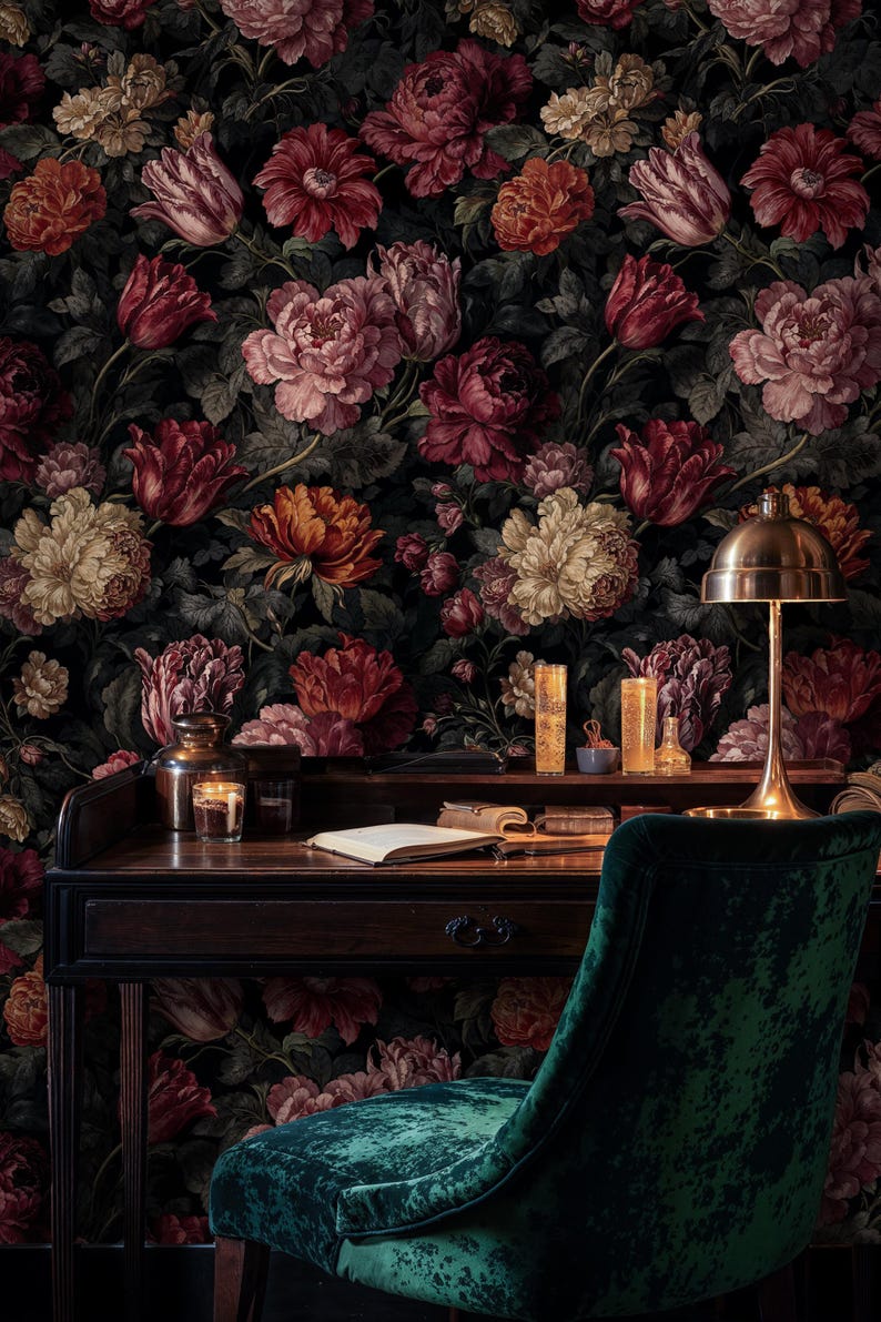 May include: A dark wooden desk with a brass lamp, books, and candles sits in front of a floral wallpaper. A green velvet chair is in the foreground. The wallpaper features large, colorful flowers in shades of red, pink, and orange.