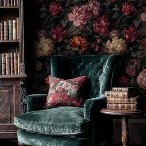 May include: A luxurious, emerald green velvet armchair with a tufted back and a floral patterned pillow. The background features a dark floral wallpaper and a wooden bookshelf filled with antique books. A small wooden side table holds a stack of old books.