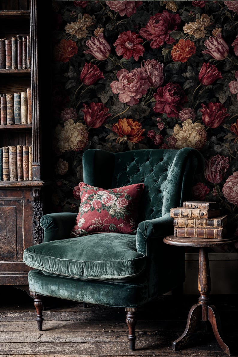 May include: A luxurious, emerald green velvet armchair with a tufted back and a floral patterned pillow. The background features a dark floral wallpaper and a wooden bookshelf filled with antique books. A small wooden side table holds a stack of old books.