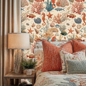 Op de afbeelding: Een slaapkamer met een bed met koraalkleurige kussens en een dekbedovertrek met patroon. Het behang heeft een zeelevenontwerp met zeepaardjes, vissen en koraal. Een houten lamp en een nachtkastje staan op de voorgrond, met een kleine plant in een pot.