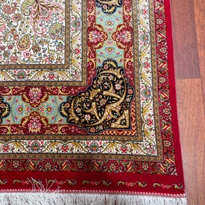 K&ouml;nnte beinhalten: Nahaufnahme eines detaillierten persischen Teppichs mit rotem Rand und wei&szlig;em Fransen. Der Teppich weist ein zentrales Design mit floralen Mustern in Rosa-, Gr&uuml;n- und Goldt&ouml;nen auf wei&szlig;em Hintergrund auf. Aufwendige Eckdesigns in Marineblau und Gold.