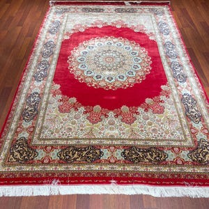 K&ouml;nnte beinhalten: Ein gro&szlig;er, rechteckiger roter Teppich mit einem komplizierten Blumenmuster und einem zentralen Medaillon-Design. Der Teppich hat einen cremefarbenen Rand mit aufwendigen Details und einen wei&szlig;en Fransen an den Kanten. Der Teppich liegt auf einem Parkettboden.