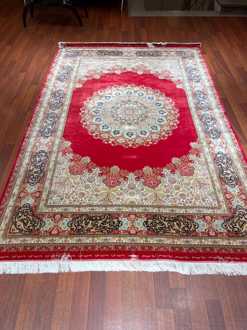 K&ouml;nnte beinhalten: Ein gro&szlig;er, rechteckiger roter Teppich mit einem komplizierten Blumenmuster und einem zentralen Medaillon-Design. Der Teppich hat einen cremefarbenen Rand mit aufwendigen Details und einen wei&szlig;en Fransen an den Kanten. Der Teppich liegt auf einem Parkettboden.