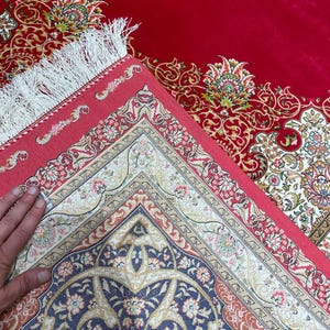 K&ouml;nnte beinhalten: Ein roter Orientteppich mit aufwendigen Blumenmustern in Creme, Blau und Pfirsich. Der Teppich hat einen detaillierten Rand und einen wei&szlig;en Fransen. Das Design ist auf der gefalteten Ecke des Teppichs sichtbar.