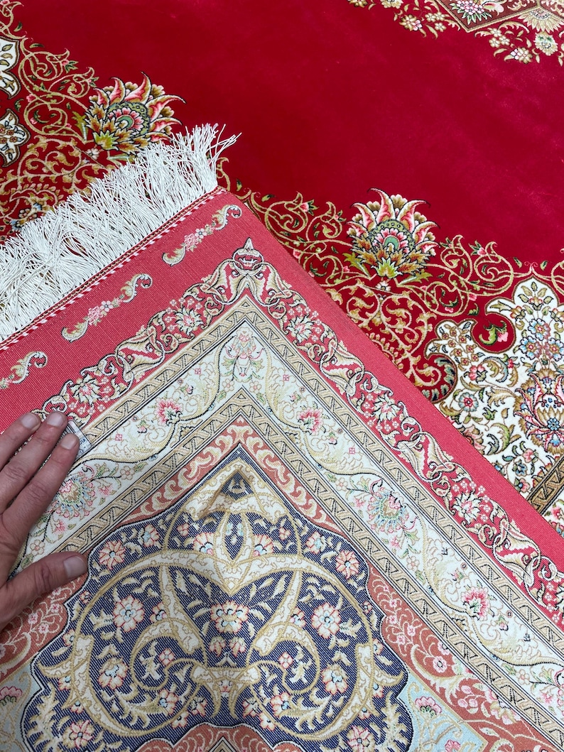 K&ouml;nnte beinhalten: Ein roter Orientteppich mit aufwendigen Blumenmustern in Creme, Blau und Pfirsich. Der Teppich hat einen detaillierten Rand und einen wei&szlig;en Fransen. Das Design ist auf der gefalteten Ecke des Teppichs sichtbar.