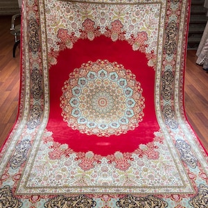 K&ouml;nnte beinhalten: Ein gro&szlig;er, rechteckiger roter Teppich mit einem komplizierten Blumen- und geometrischen Muster. Der Teppich hat ein zentrales Medaillon in Blau-, Gr&uuml;n- und Goldt&ouml;nen, umgeben von einem detaillierten Rand mit &auml;hnlichen Farben und Designs. Der Teppich hat einen Fransen.