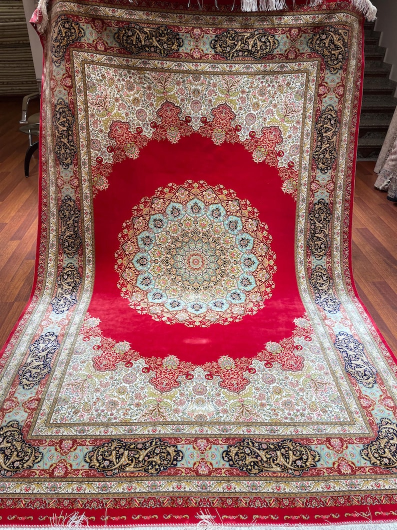K&ouml;nnte beinhalten: Ein gro&szlig;er, rechteckiger roter Teppich mit einem komplizierten Blumen- und geometrischen Muster. Der Teppich hat ein zentrales Medaillon in Blau-, Gr&uuml;n- und Goldt&ouml;nen, umgeben von einem detaillierten Rand mit &auml;hnlichen Farben und Designs. Der Teppich hat einen Fransen.