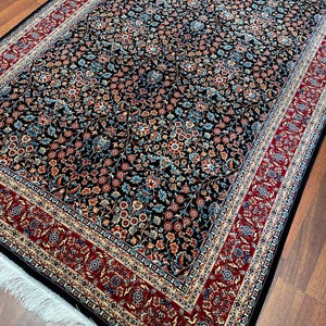 May include: A rectangular black rug with an intricate floral pattern in shades of red, blue, and beige. The rug has a red border with a detailed design and white fringe on one end. The rug is placed on a wooden floor.