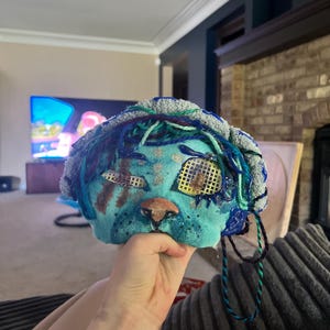Op de afbeelding: Een handgemaakt turquoise masker met een textuurrijke grijze rand, blauwe garenaccenten en een oogbedekking met een rasterpatroon. Het masker heeft een bruine neus, een klein zilverkleurig ringdetail en een pleisterachtig stuk over het rechteroog.
