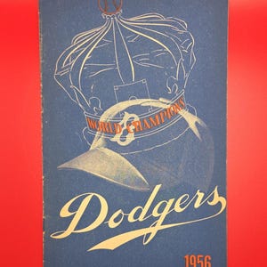 May include: A vintage Dodgers baseball program from 1956, featuring a blue cover with a baseball, crown, and the words "World Champions." The team name "Dodgers" is in a stylized script, with the year 1956 in orange.