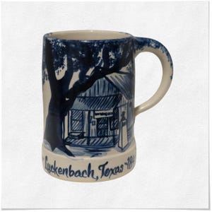 May include: A ceramic mug with a blue and white illustration of a building and tree. The mug has a handle and the words "Luckenbach, Texas 1849" are written on the bottom. The mug is cylindrical and has a slightly flared base.