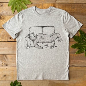 Puede incluir: Una camiseta gris con una ilustración en blanco y negro de un oso tumbado en un sofá. El oso sostiene un cuenco de fideos en sus patas.