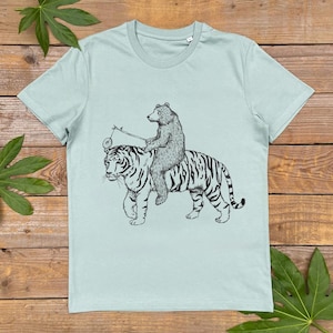 May include: A light blue t-shirt with a black and white illustration of a bear riding a tiger. The bear is holding a stick with a ring on the end.