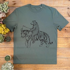 May include: A green t-shirt with a black and white illustration of a bear riding a tiger. The bear is holding a stick with a circle on the end.